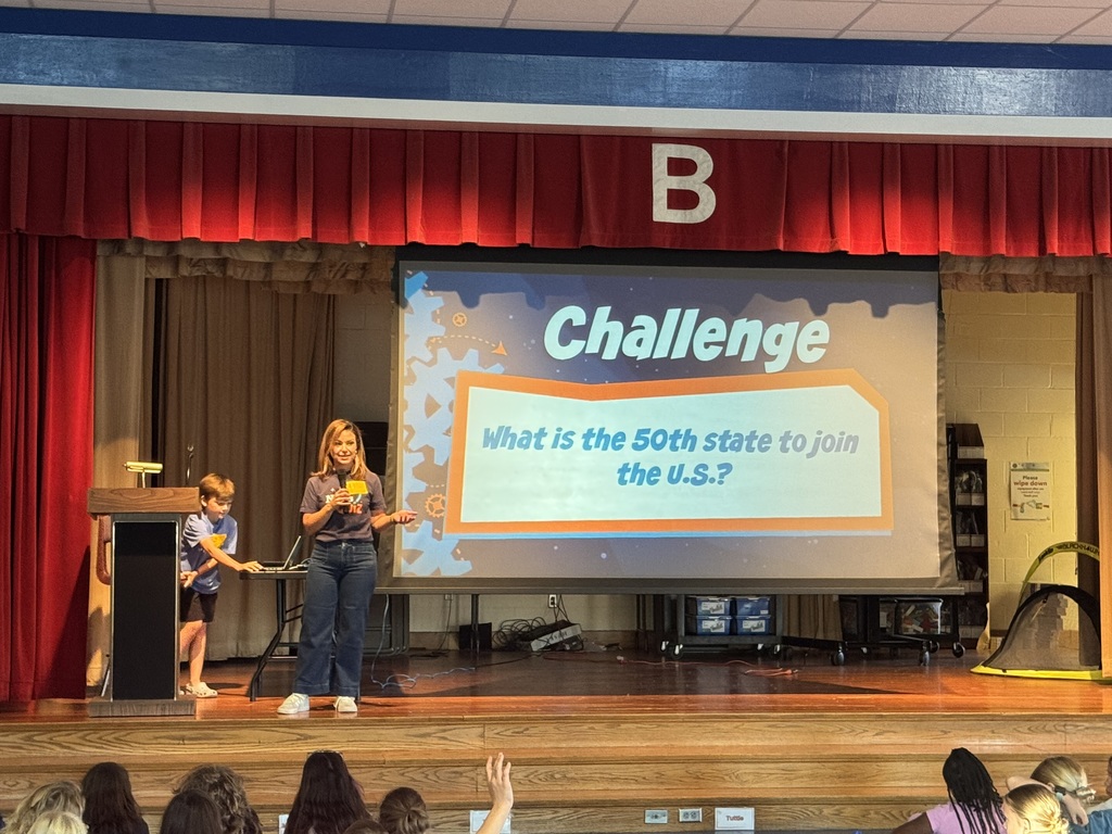 An adult stands on a stage and speaks into a microphone while a projector screen behind her reads: "Challenge: What is the 50th state to join the U.S.?"