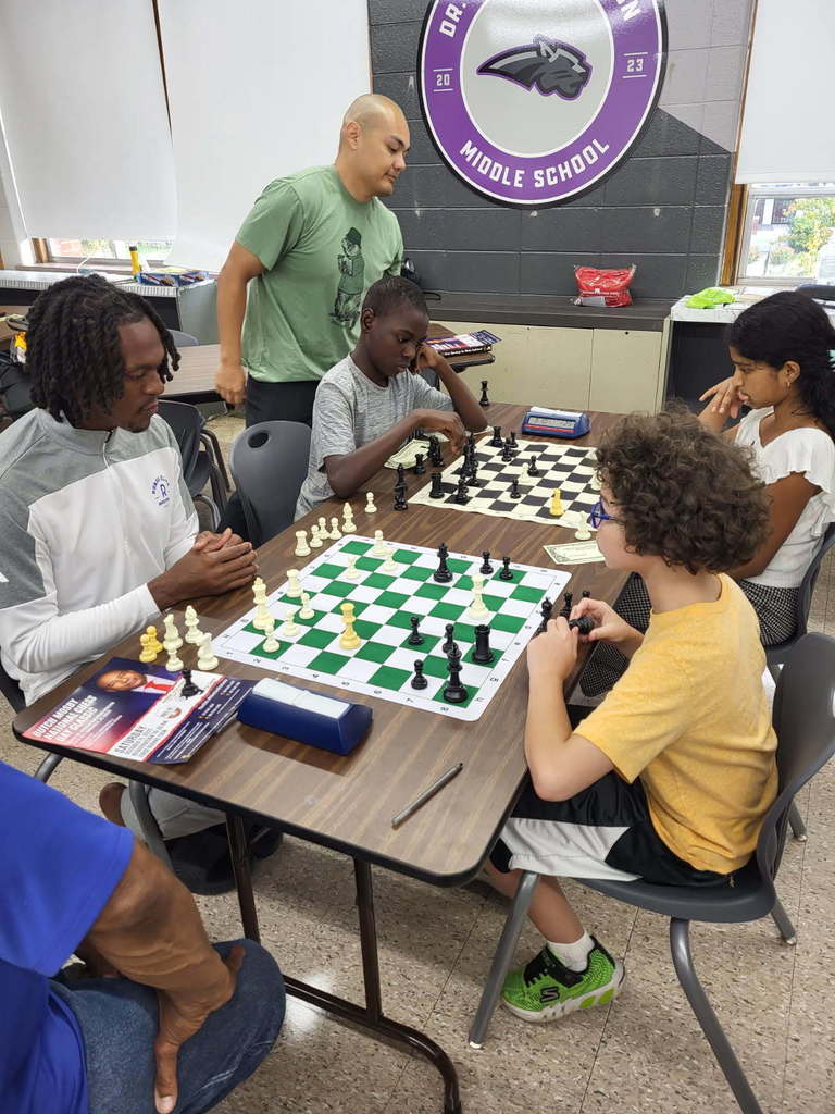 An adult. plays a student in chess. Two other students play a chess game nearby.