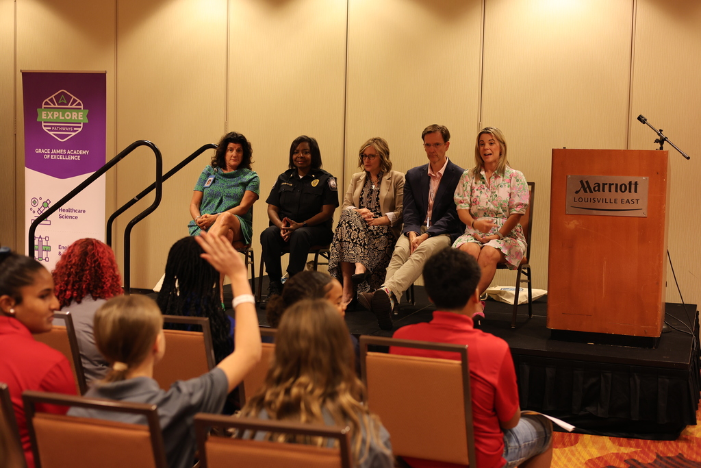 Five adults sit on a stage while students sit in chairs in the audience.