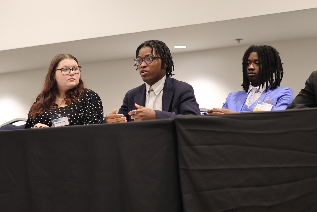 Three students sit at a table while one speaks.