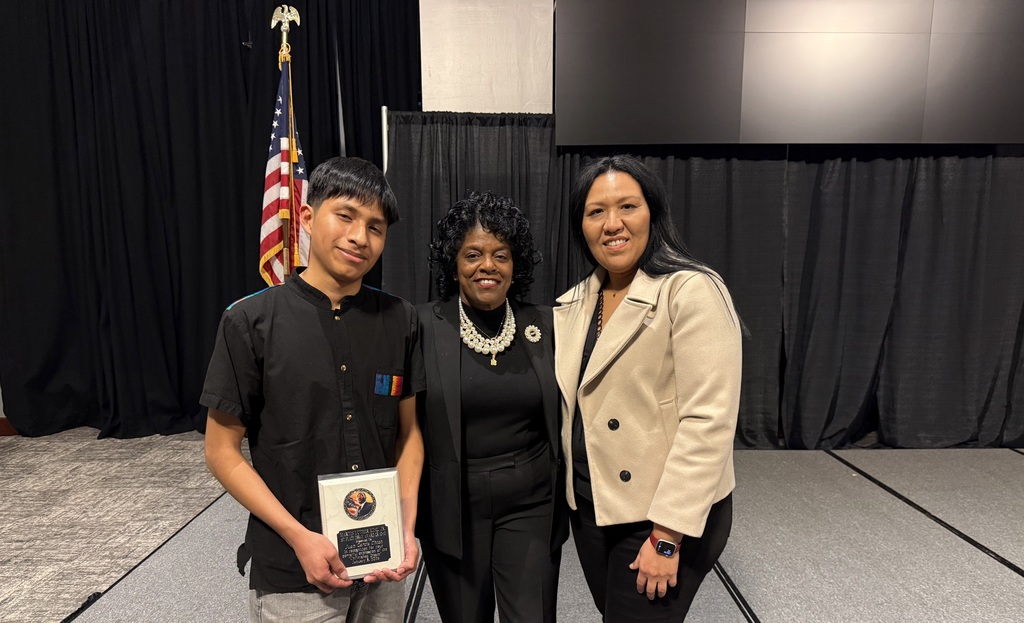 A student holds a plaque and poses for a picture with two adults.