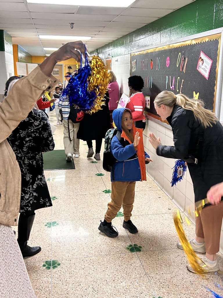 A student does a high five with a Mornings with Mom volunteer as other volunteers stand and cheer in the school hallway.