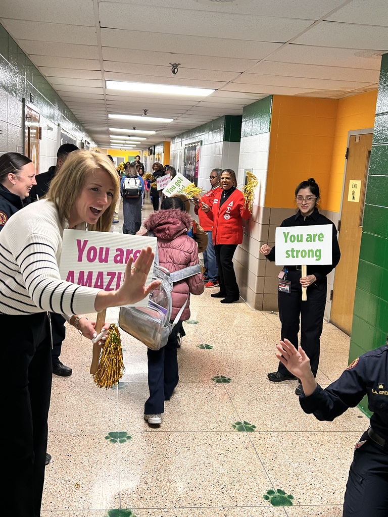 A student does a high five with a Mornings with Mom volunteer as other volunteers stand and cheer in the school hallway.