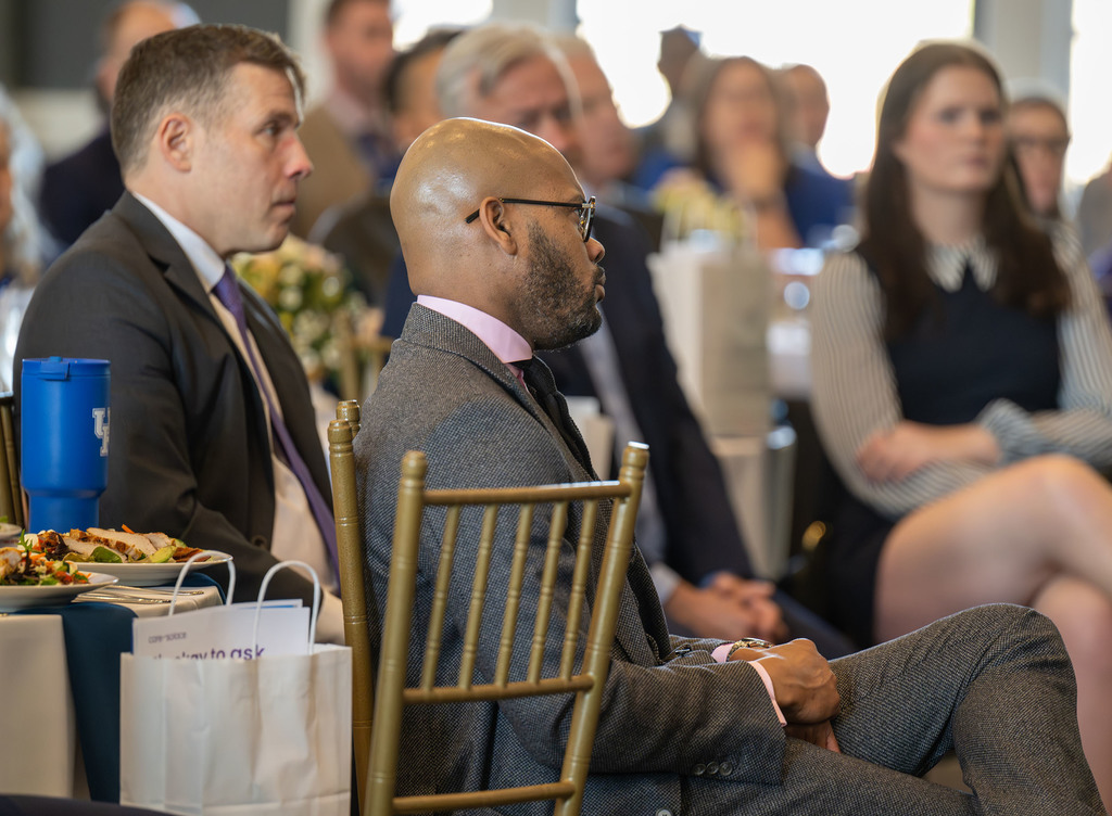 Adults sit in chairs and listen to a speech.