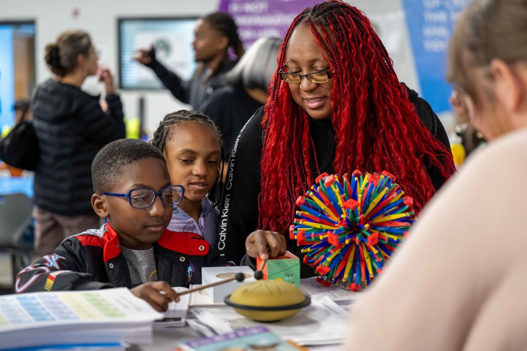 A child hits an object with two mallets while adults and another child watch. 