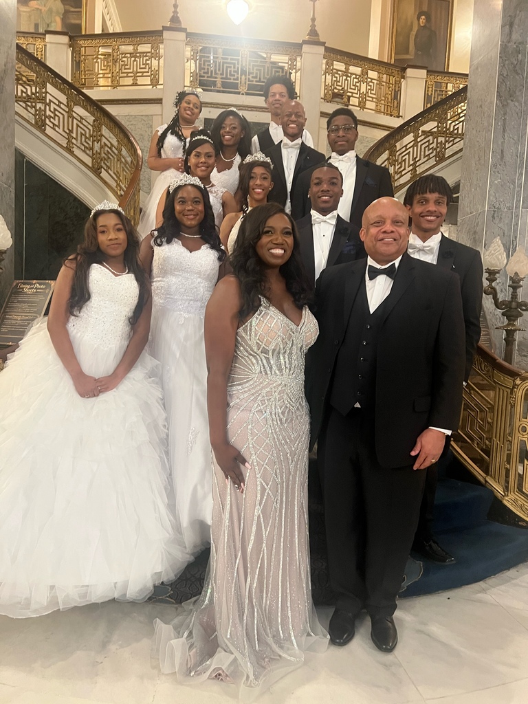 Thirteen individuals wearing formal wear pose for a photo and smile. 
