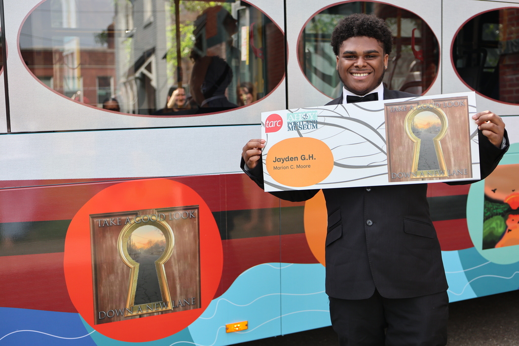 A student holds a copy of his design and stands by that design on the TARC bus. 