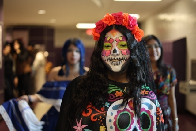 A young person dressed in bright Day of the Dead sugar skull makeup and a floral headband stands in a school hallway,