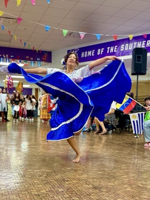 A female student dances barefoot in a school cafeteria or gym, performing a traditional Latin dance.