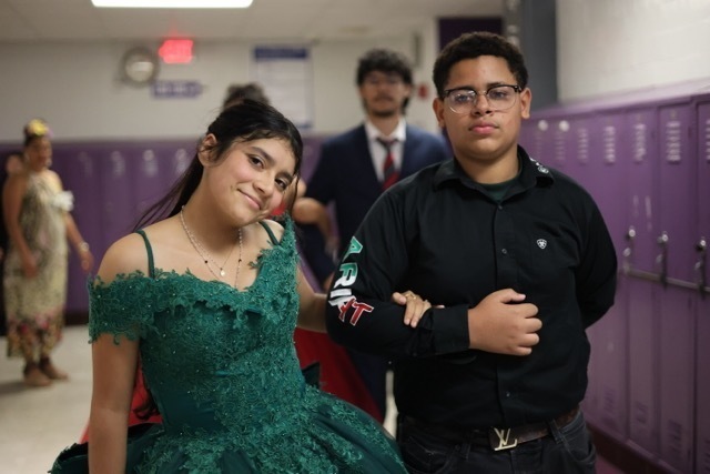 Two students, a girl and a boy, stand posing together in a school hallway with purple lockers. The girl wears a voluminous emerald green off-the-shoulder dress, and the boy wears a black button-down shirt. Other