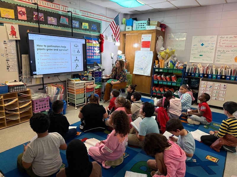 A teacher sits in the front of a classroom while students sit on a carpet.