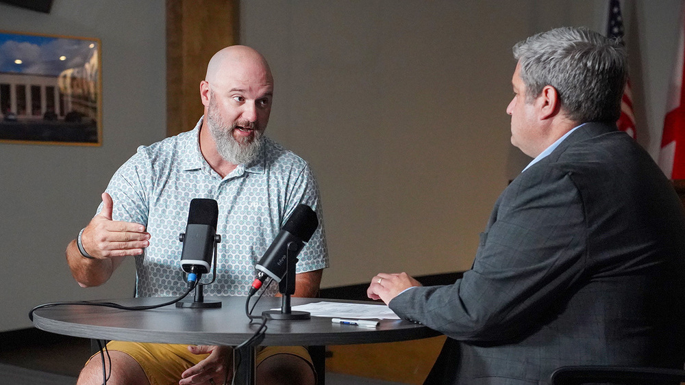 Clay-Chalkville High School football coach Stuart Floyd speaks into a microphone while looking at a podcast host. They both sit at a table in a room.