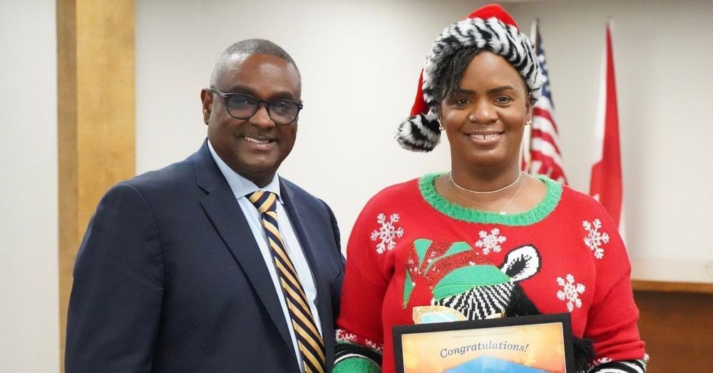Image is of JEFCOED Superintendent Dr. Walter Gonsoulin and Monica Spencer standing together and smiling for a photo in the Jefferson County Schools Board Room. Dr. Gonsoulin is wearing a suit and Monica Spencer is wearing a Santa hat trimmed with Zebra print and a holiday sweater with a zebra on it. She holds a JEFCOED Strong Award.