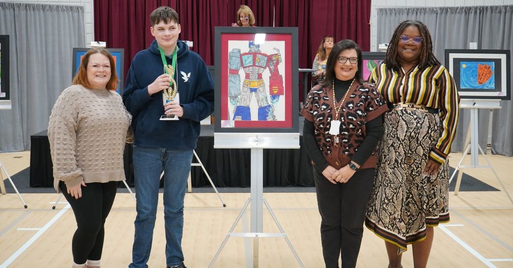 A high school student and four women pose together for a photo next to art created by the student. The student has a medal around his neck.