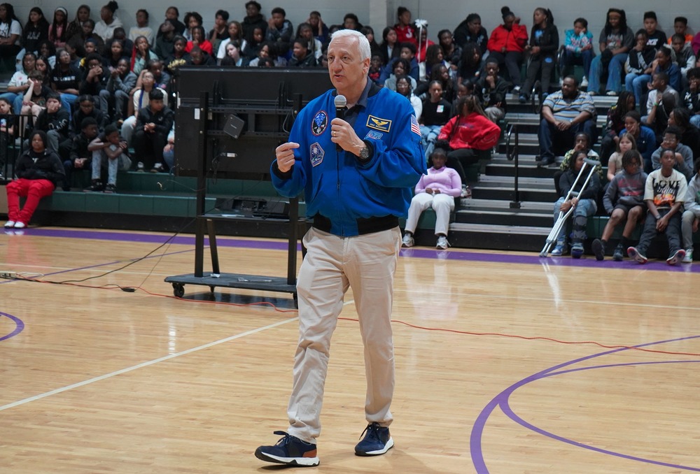 Former NASA Astronaut Mike Massimino speaks into a microphone while talking to students at Minor Middle School. He stands on the gym floor while students sit in bleachers.