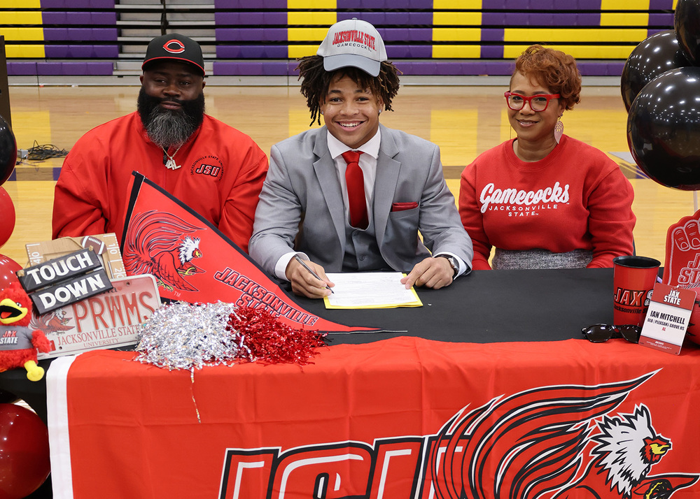Student Signs a letter of intent to continue his athletic career with Jacksonville State University. He is surrounded by his family and is smiling brightly. 