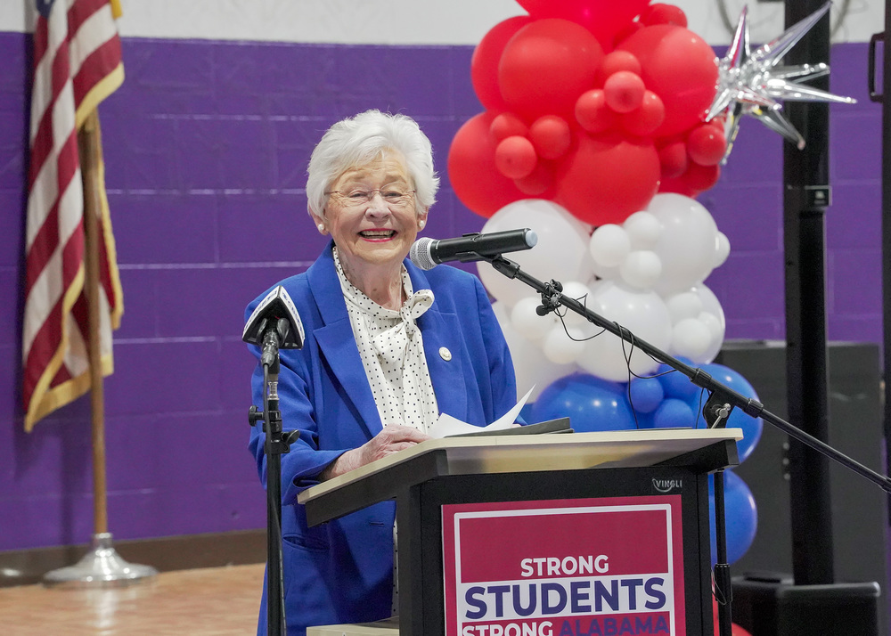 Gov. Kay Ivey smiles while standing at a podium in a school gym.
