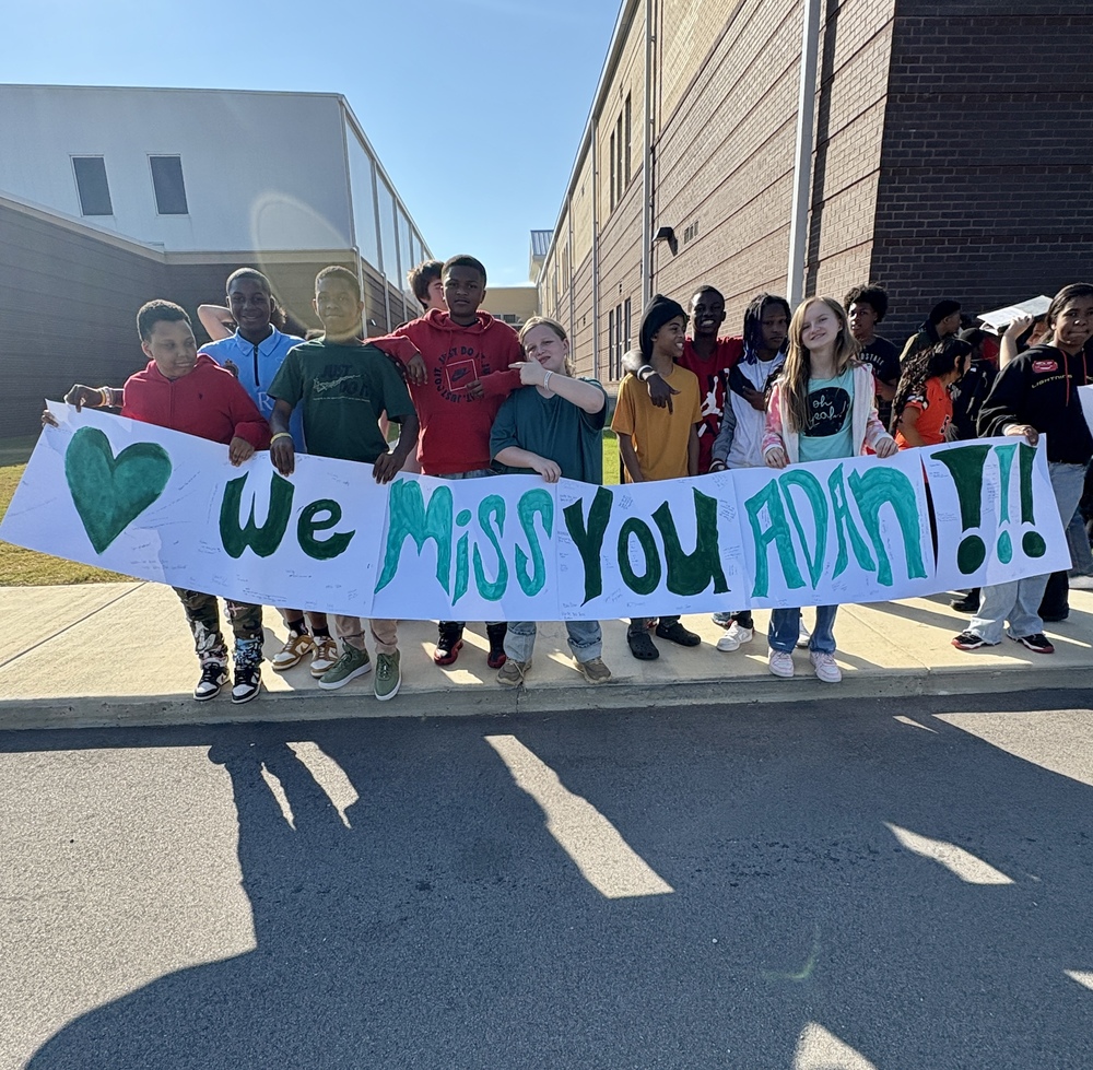 A group of students stand outside a school, holding a sign that says "We miss you Adan!!!"