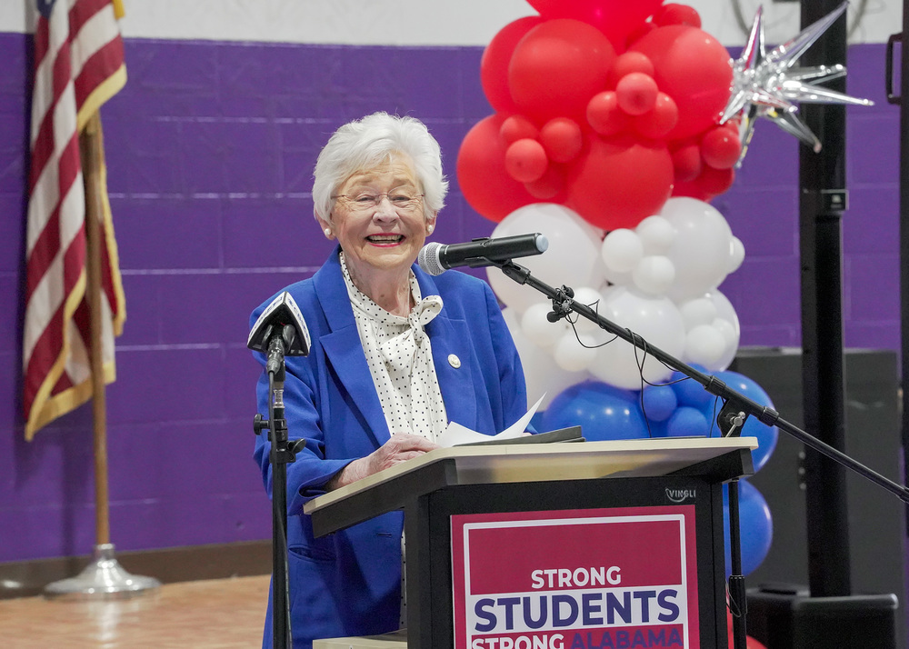 Gov. Kay Ivey smiles while standing at a podium in a school gym.