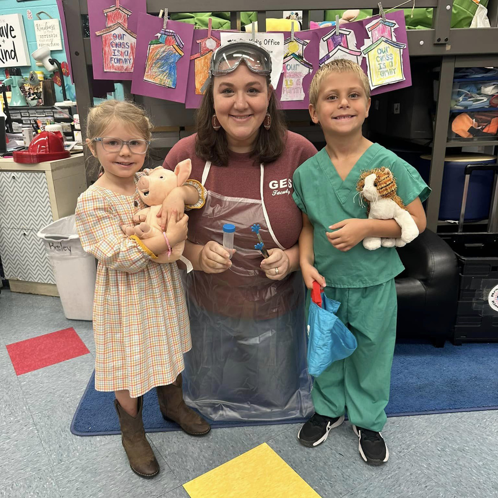 Two students standing next to a teacher in class. They are holding stuffed animals.