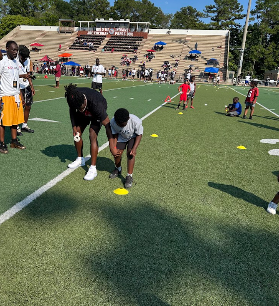 Many people stand on a football field in an outdoor stadium. At the center of the photo, a man helps a young boy while he does football drills.