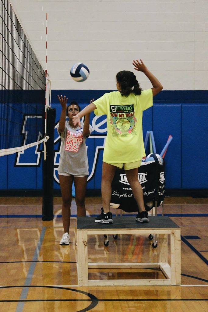 A teen girl tosses a volleyball into the air as a young girl stands on an elevated platform, trying to hit the ball.