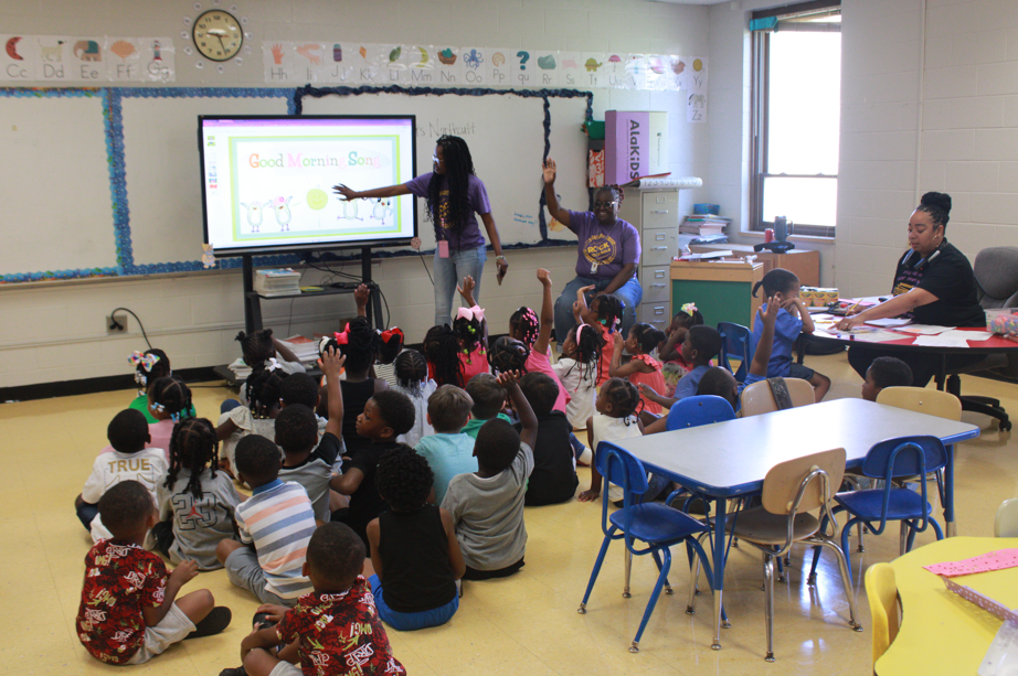A group of students sit on the ground while teachers speak to them from the front of a classroom.