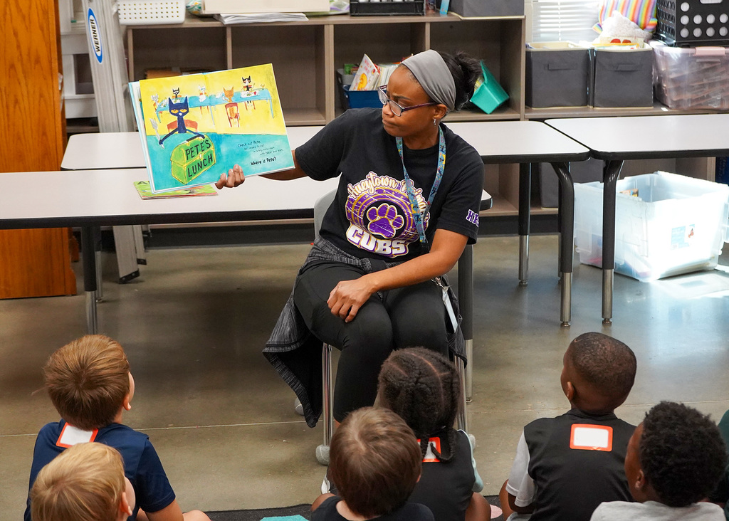 A teacher holds a book up while sitting in a chair and reading to students