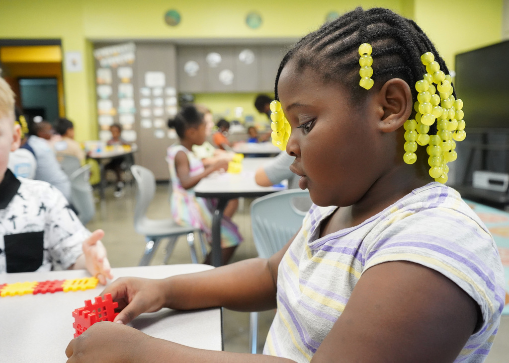 A little girl sits at a table in a classroom and holds a toy