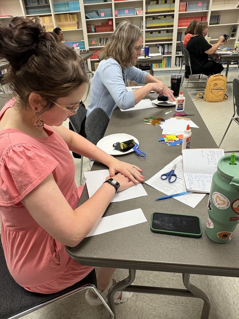 Women sit at tables in a room. One woman is pressing down on a piece of paper. Another woman is placing a paint brush into paint on a paper plate.