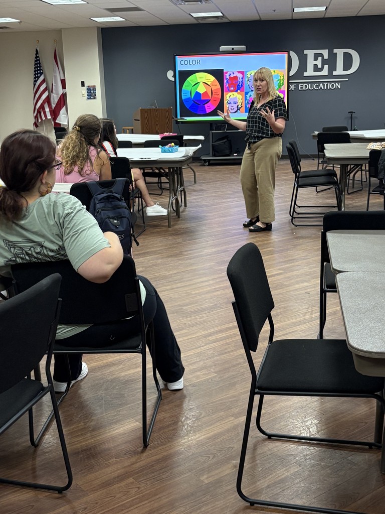 Woman stands in front of room teaching a lesson to other adults.