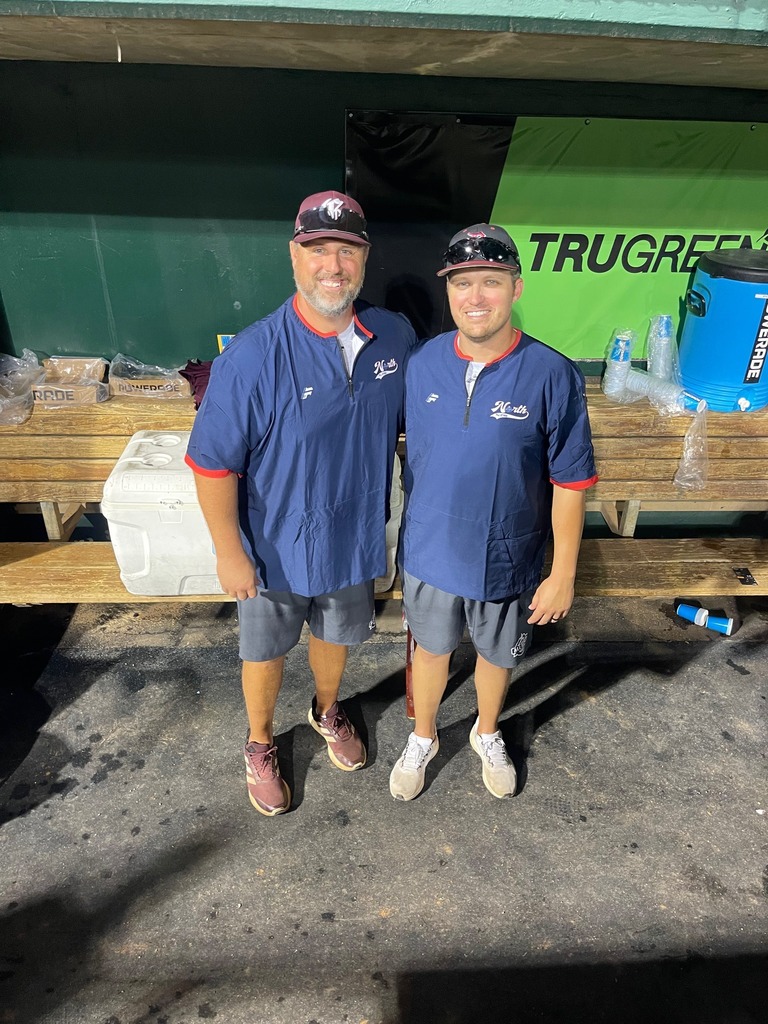 Two men wearing athletic clothes stand in a dugout, smiling at camera.