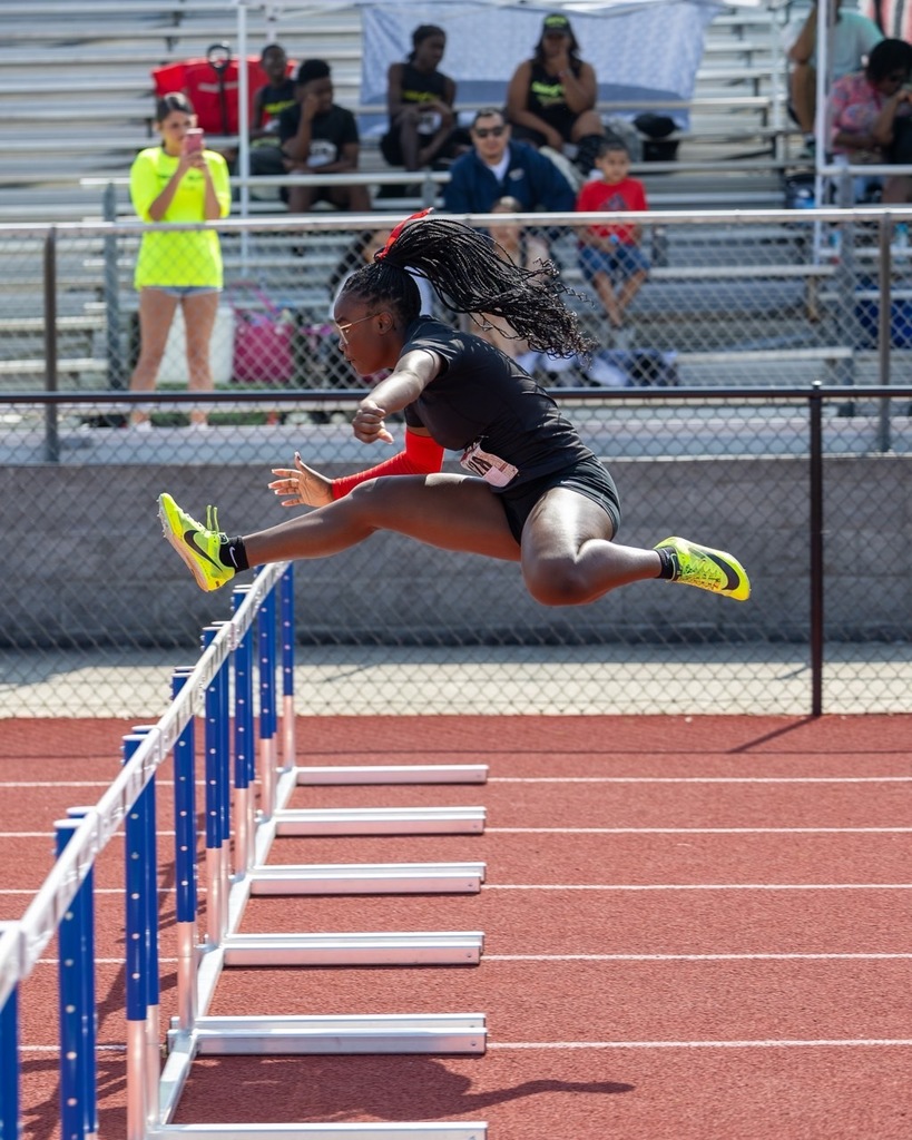 A girl jumps over a hurdle on an outdoor track. People in the background sit on bleachers.