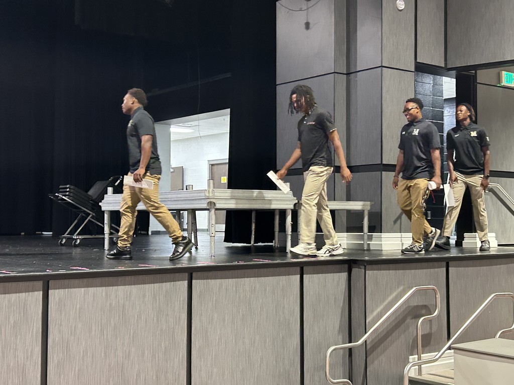 Four students wearing matching polo shirts and khaki pants walk onto a stage.