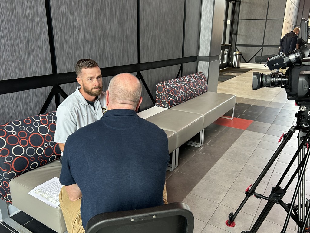 Two men sit in a room, facing each other. A camera on a tripod sits next to the two men.