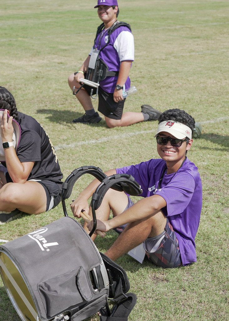 Three students sit on grass, two are smiling at the camera. A drum sits on the ground next to one of the students.