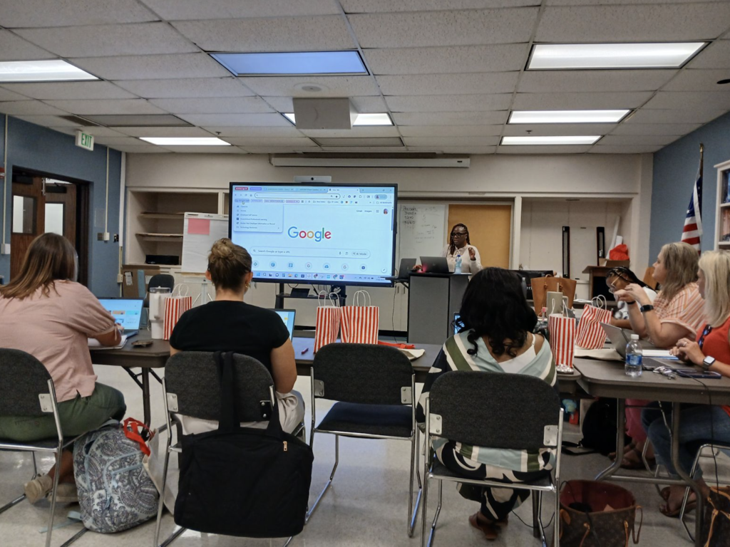 A woman stands next to a board, giving a presentation to a room of adults.