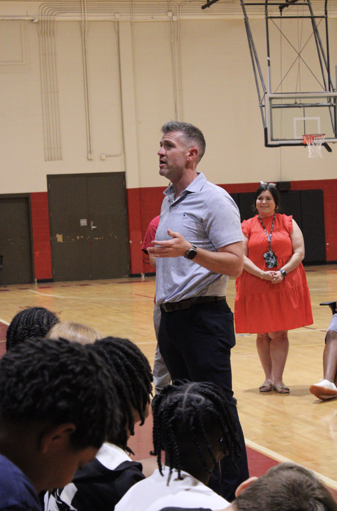 A man stands in a gym, speaking to a group of students