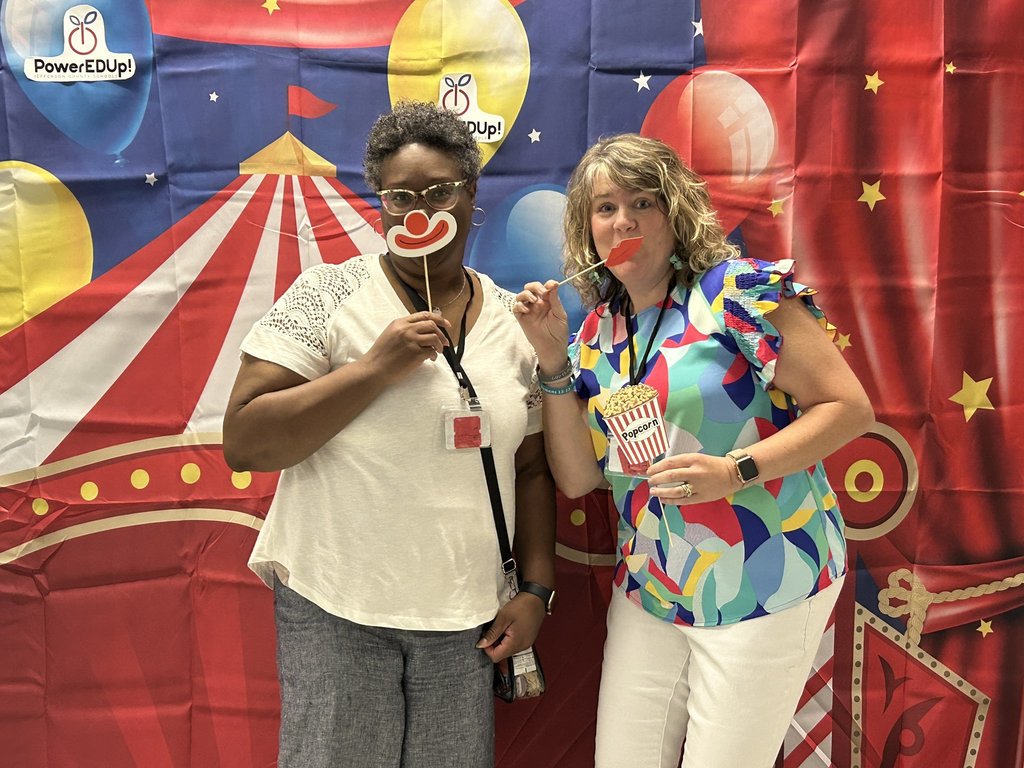 Two women stand in front of a circus themed photo backdrop. The women hold photo props over their faces.