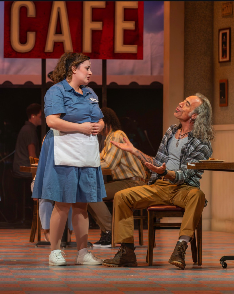 A man and a woman stand on a stage and talk to each other. Two people sit at a table in the background behind the man and woman. The set is designed to look like a diner.