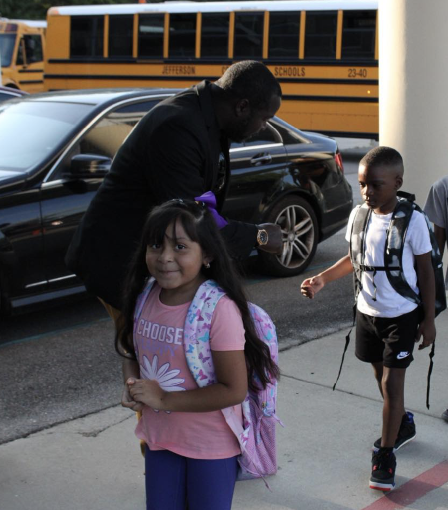 A little girl wearing a backpack smiles while walking on a sidewalk. In the background, a man greets a young boy. Behind them are school buses and cars in a parking lot.