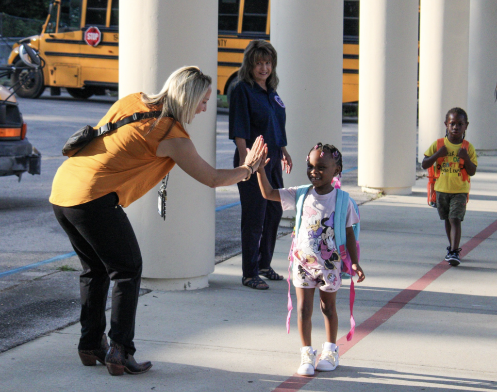 An adult gives a young student a high-five as she walks on a sidewalk. Another adult and a child stand in the background. 