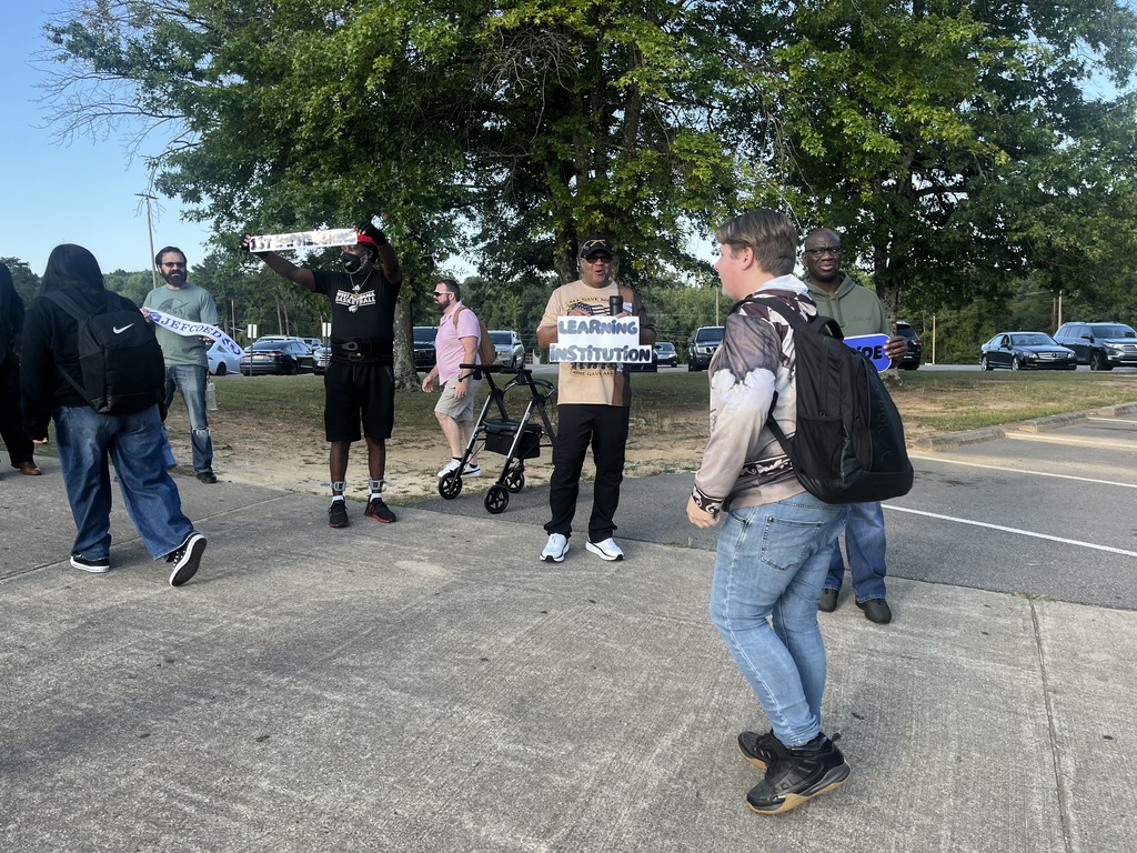 High school students walk on a sidewalk while adults hold school related signs and smile