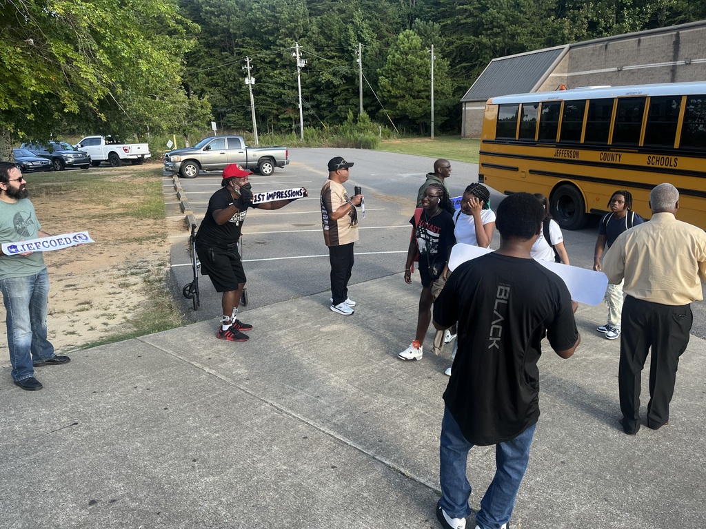 Students walk on a sidewalk after getting off a bus. They're surrounded by adults holding encouraging, school-related signs.