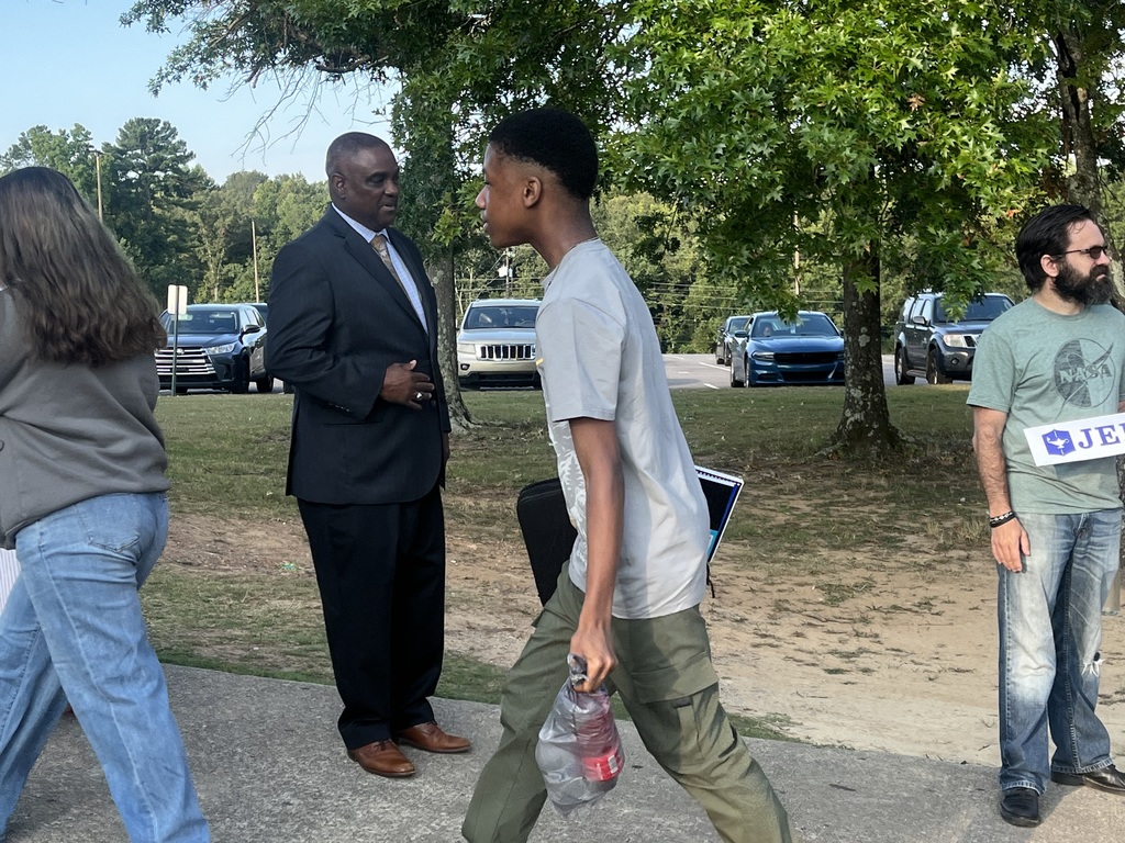 Students walk on a sidewalk after getting off a bus. Adults stand next to them, greeting the students with smiles and school-related signs.