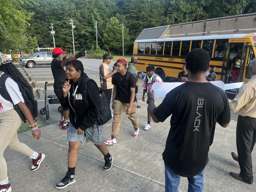 Students walk on a sidewalk after getting off a bus. They're surrounded by adults who are holding signs.