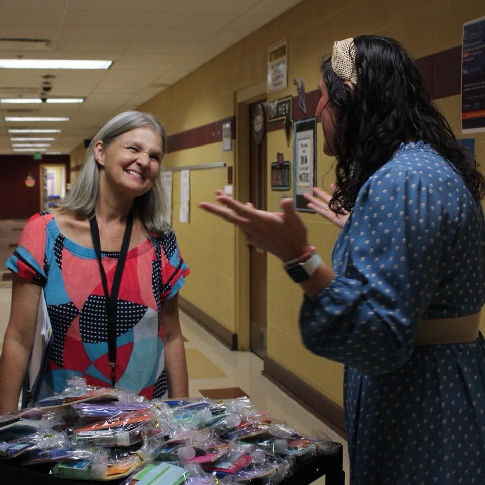 Two women stand next to a cart with goodie bags on it. The two women face each other, talking.