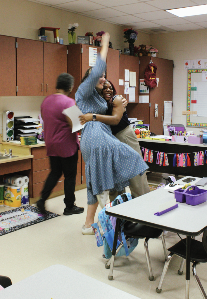 Two women hug while standing in a classroom. Another woman walks behind them.