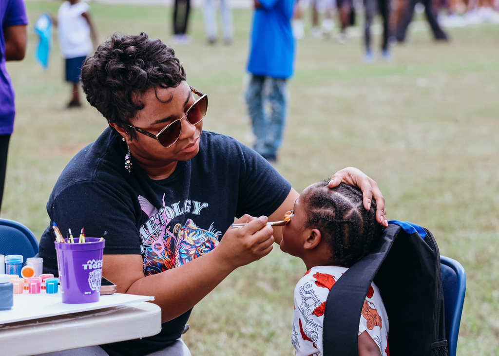 An adult and child sit on chairs outside in a grassy field. The adult paints the child's face.