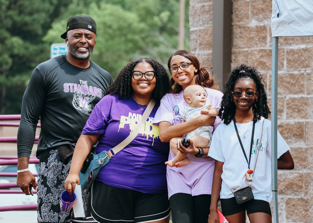 Four people stand outside and smile. One of them, a woman, holds a baby.